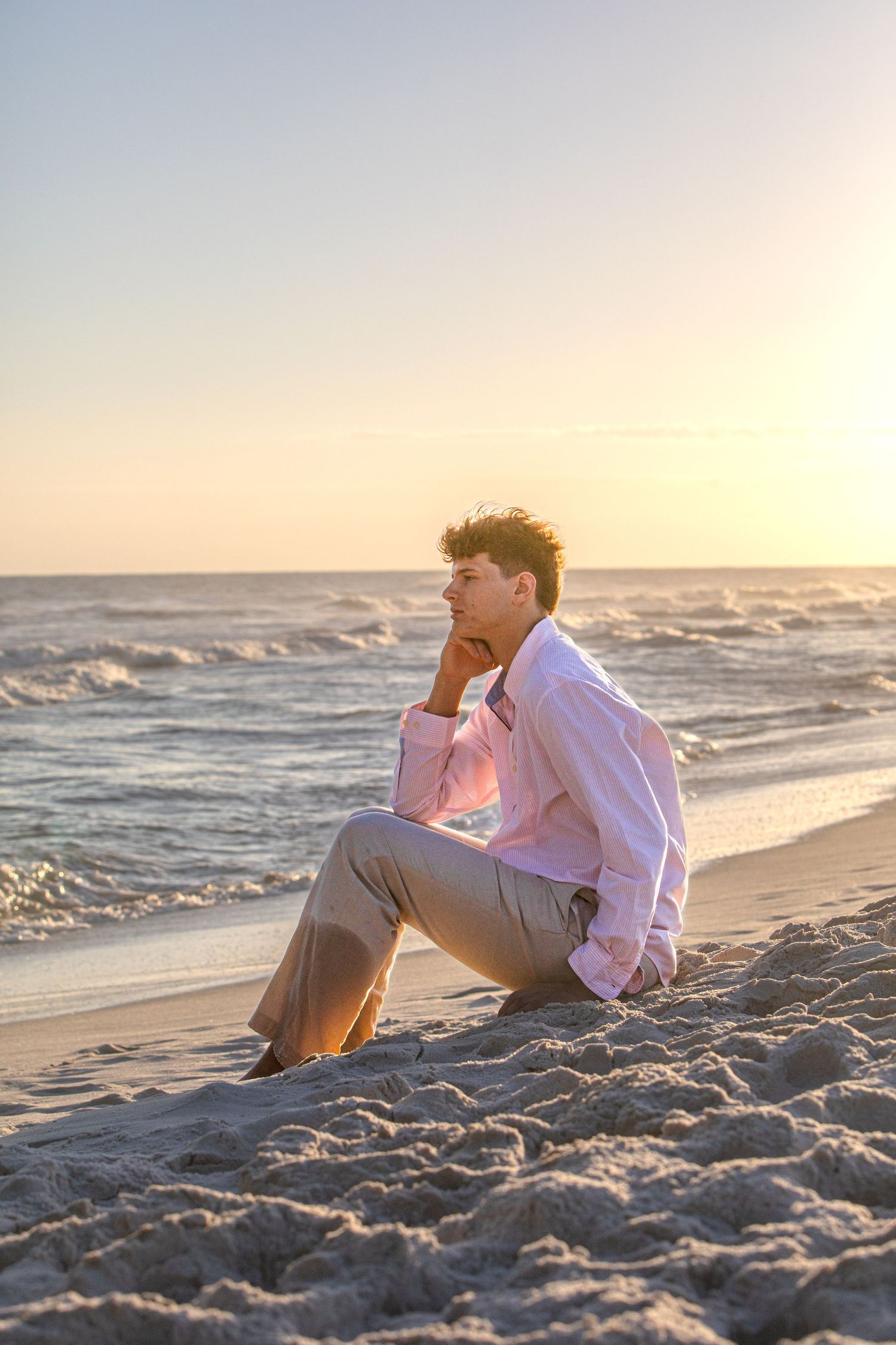A person in a pink shirt and khaki pants sits thoughtfully on the beach at sunset.