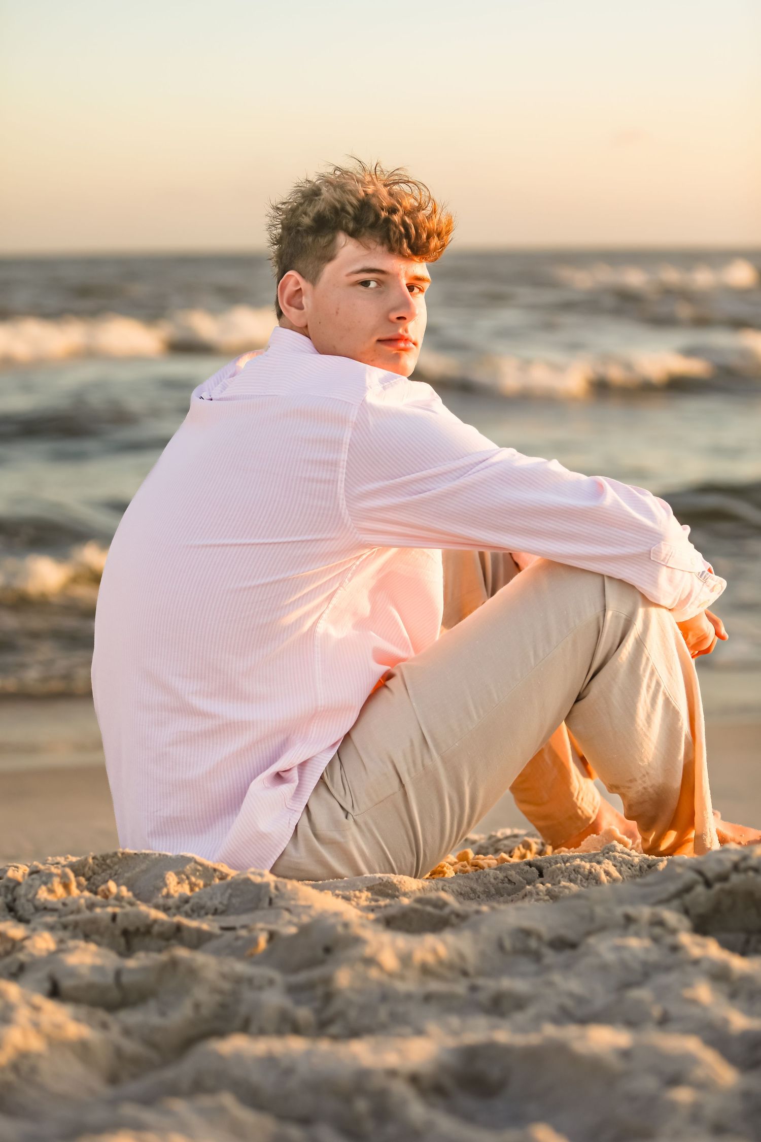 Golden hour beach portrait of person in white button-down shirt sitting on sand.