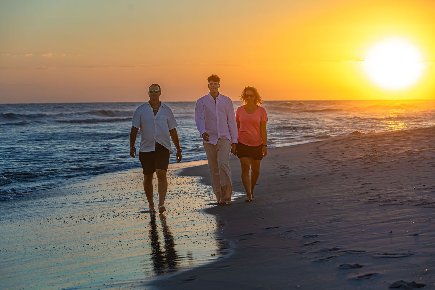 Three people walking along beach shoreline during vibrant orange sunset.