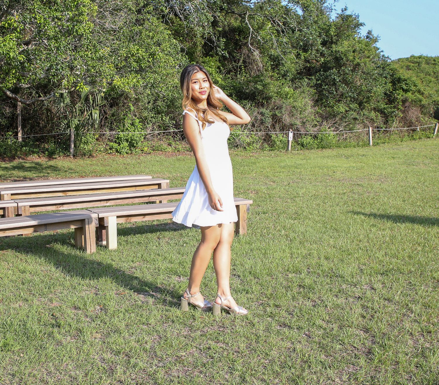 A person in a white dress poses near wooden benches in a grassy park setting.