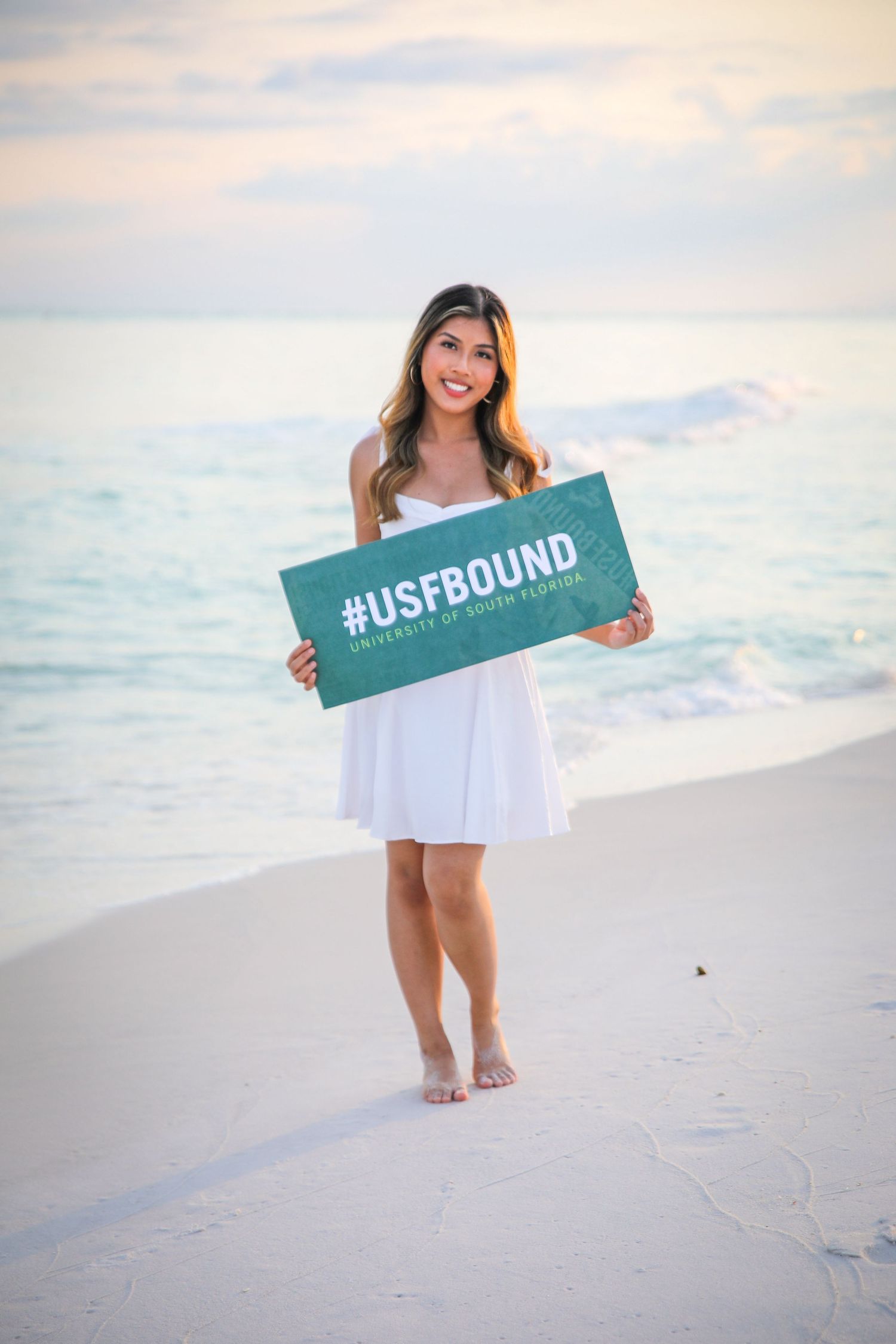 A person holds a #USFBOUND sign while standing on a beach at sunset.