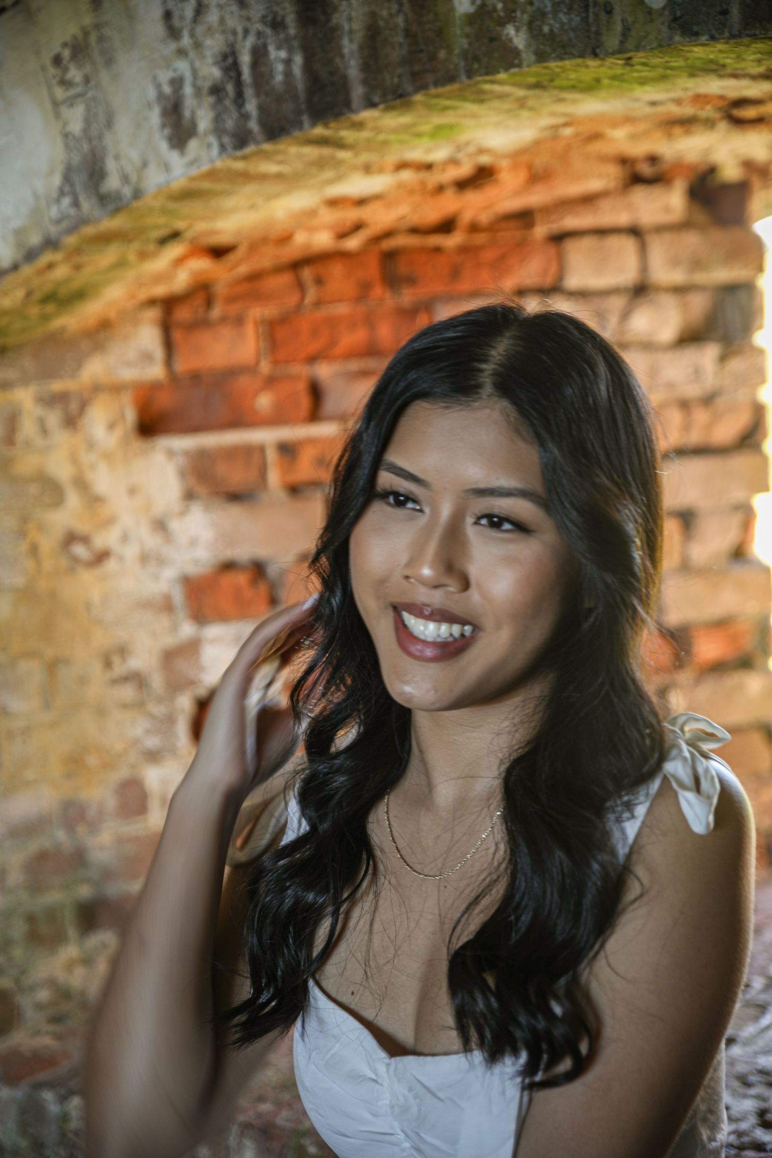 A person in a white dress poses against a weathered brick wall with natural lighting.