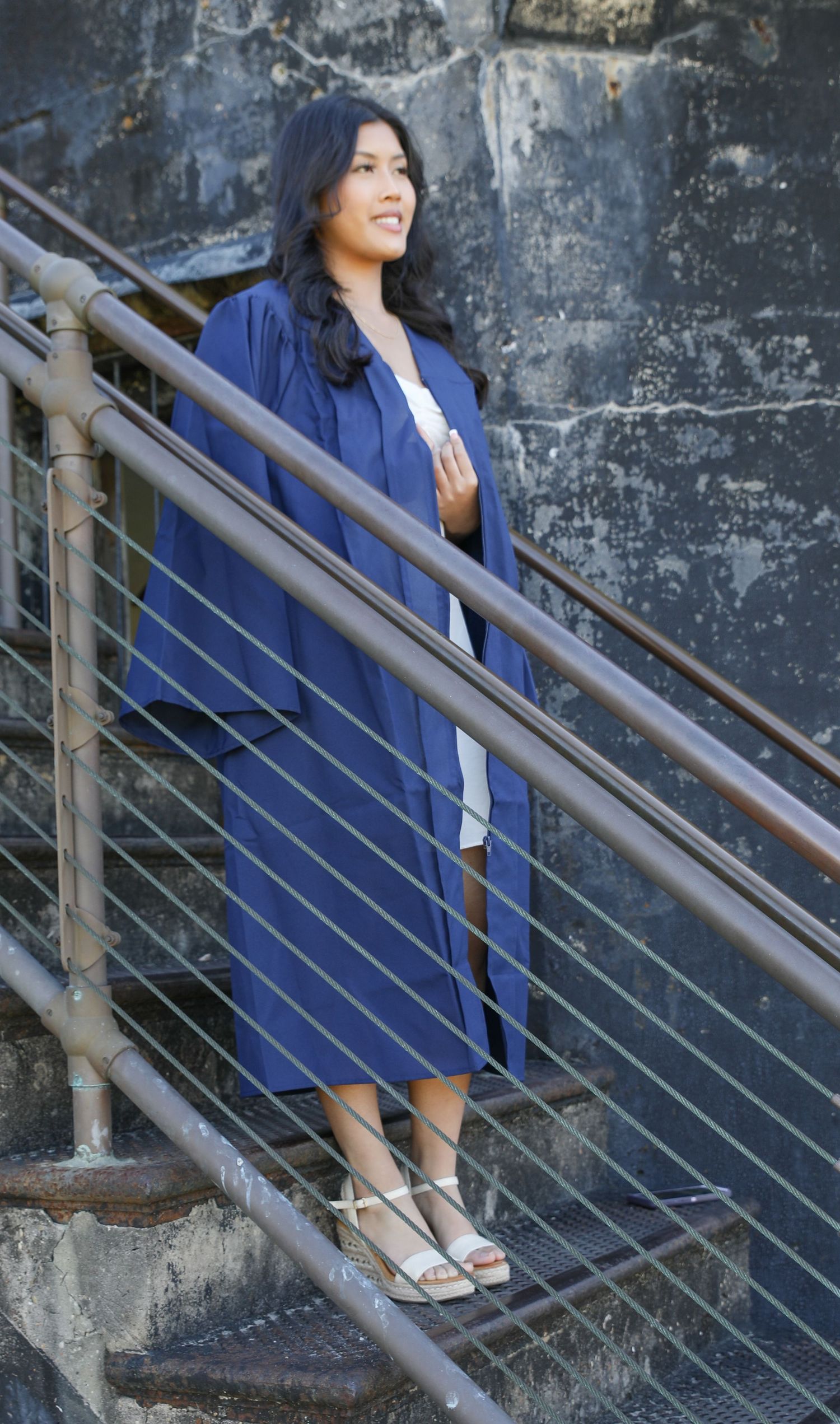 A person in a navy blue dress poses on concrete stairs against an industrial wall background.