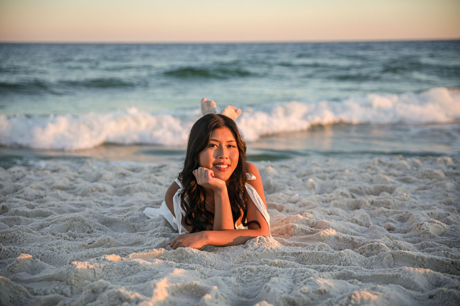 A person lies on white sand at sunset with ocean waves in the background.