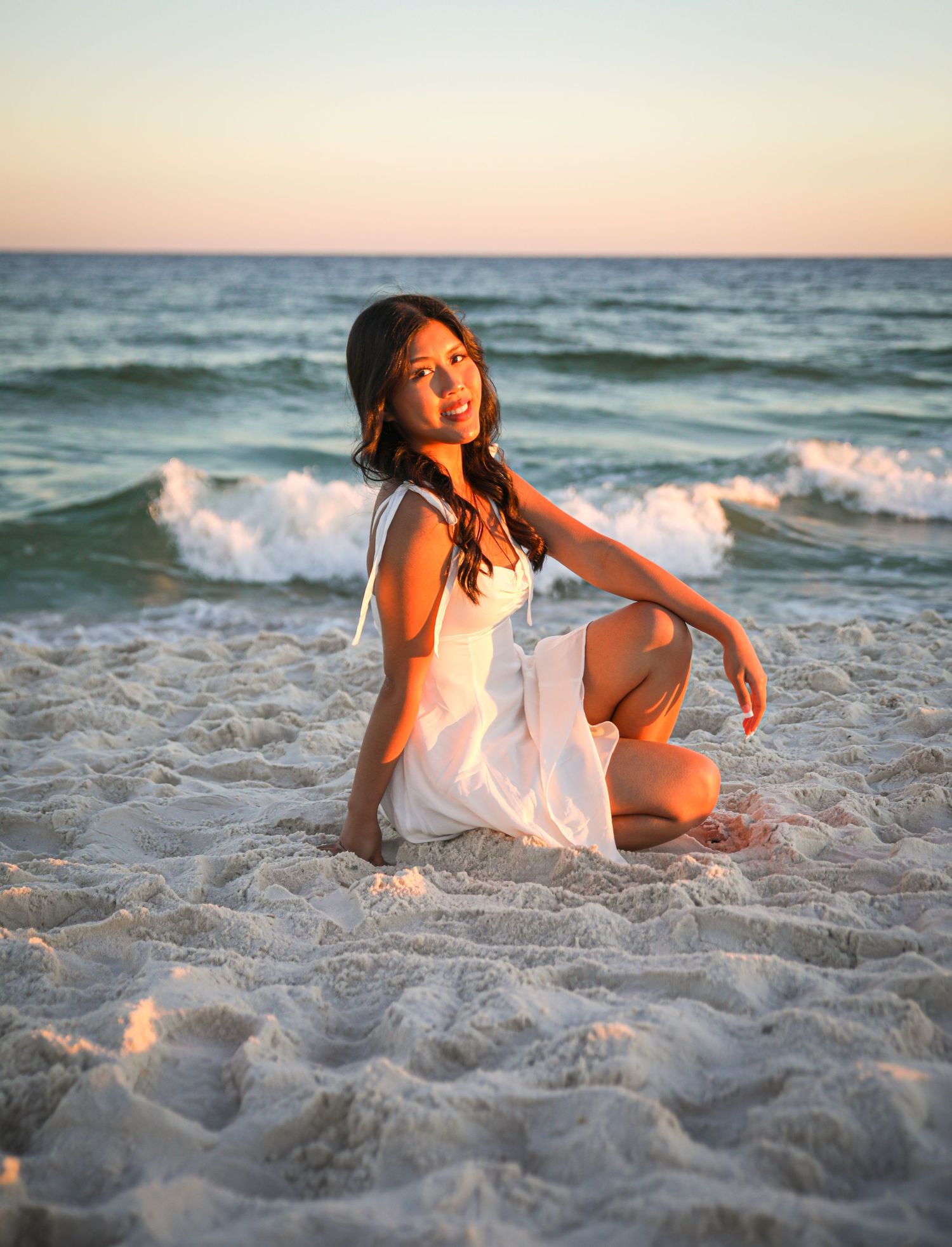 A person in white dress poses playfully on beach sand during golden hour.