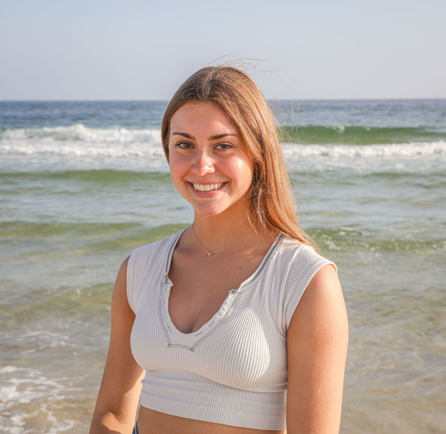 A person in a white crop top smiles warmly against an ocean backdrop on a sunny day.