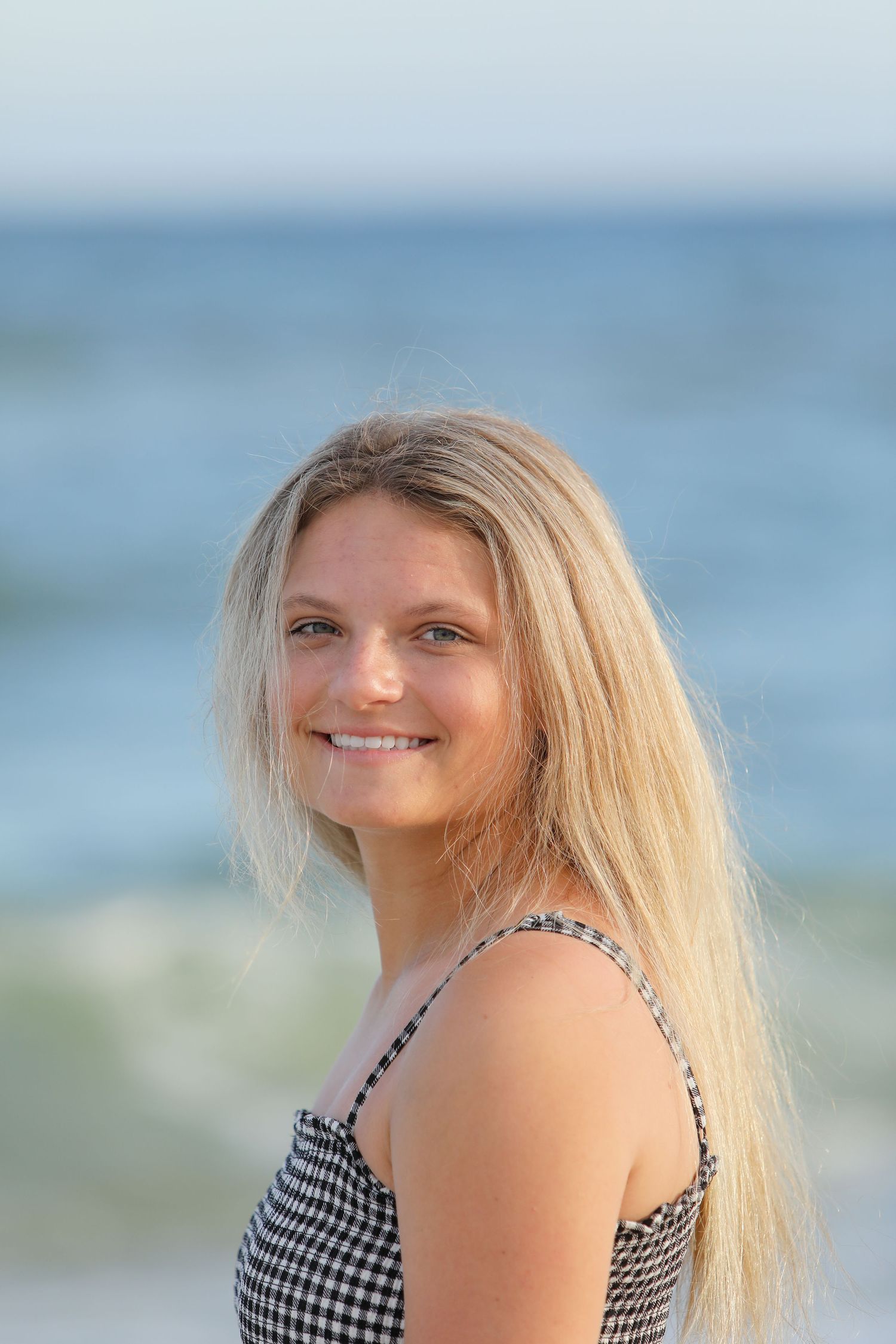 A portrait of a person in a gingham top smiling by the ocean during golden hour.