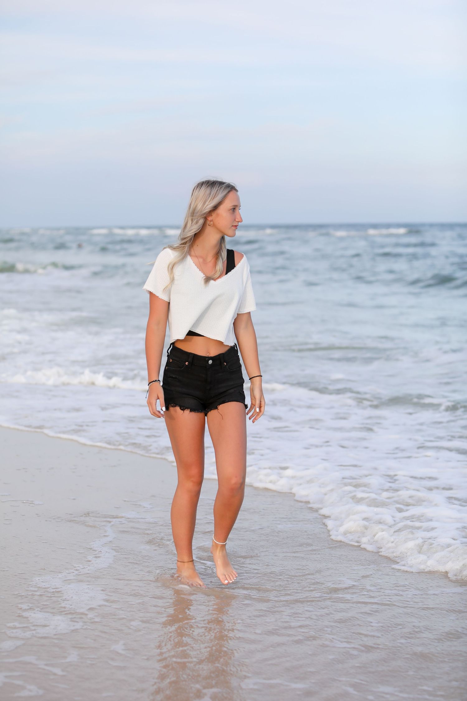 A beachgoer in a white crop top and black shorts walks along the ocean shoreline.