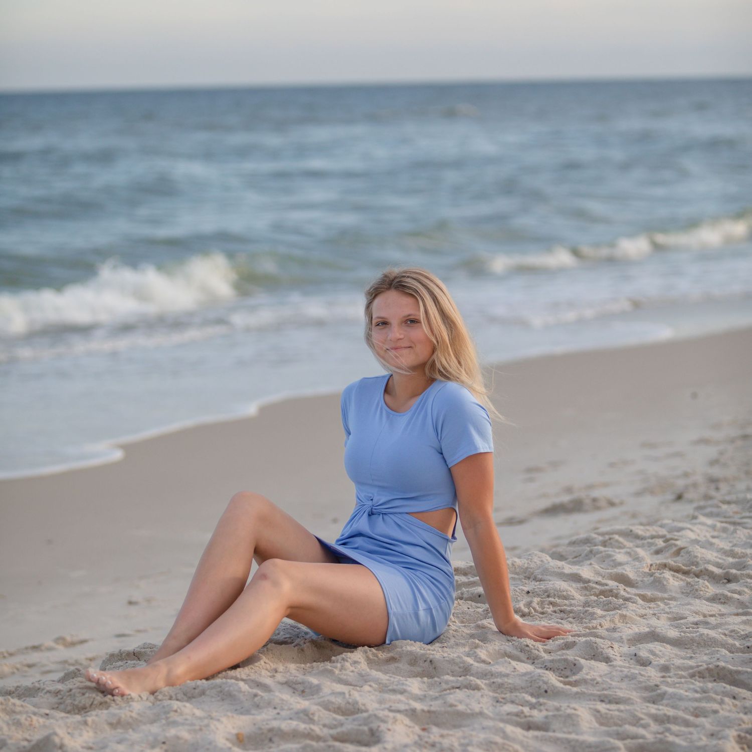 A person in a blue dress sits on the beach at sunset with waves in the background.