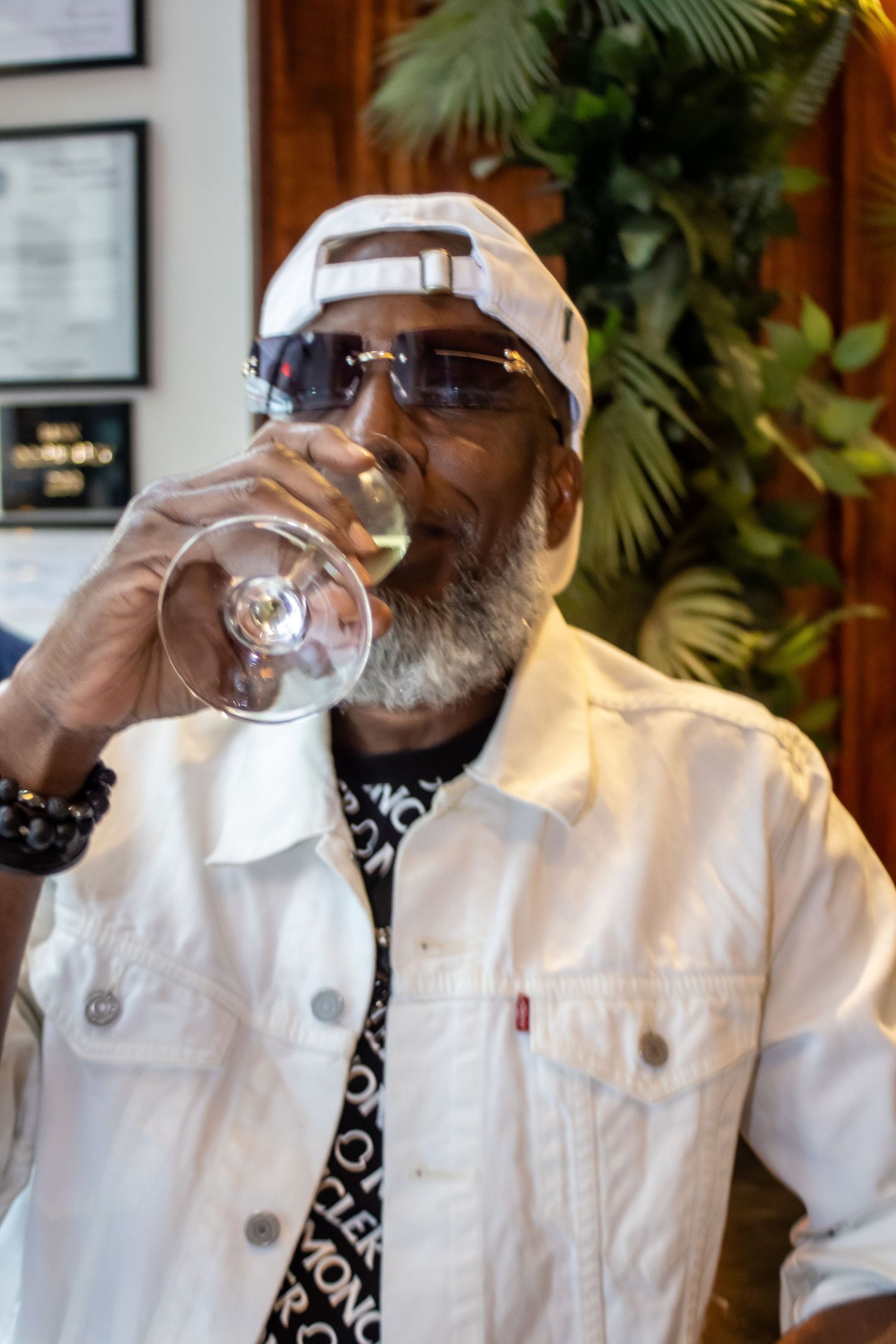 Person in white denim jacket and baseball cap sipping from a wine glass.