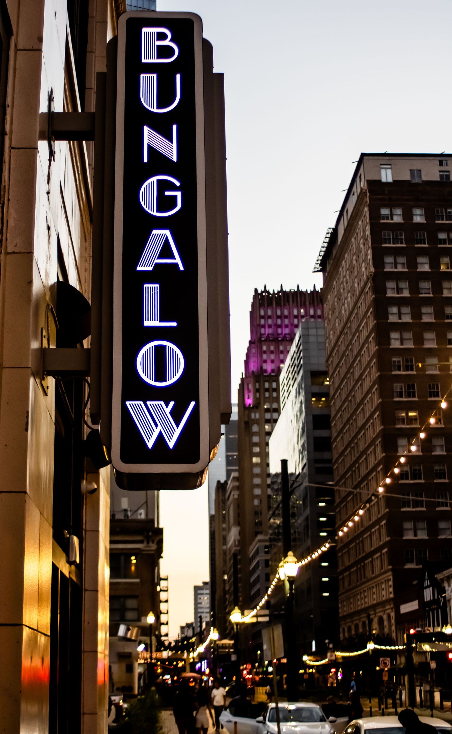 Illuminated Bungalow neon sign on city building during dusk with urban skyline.