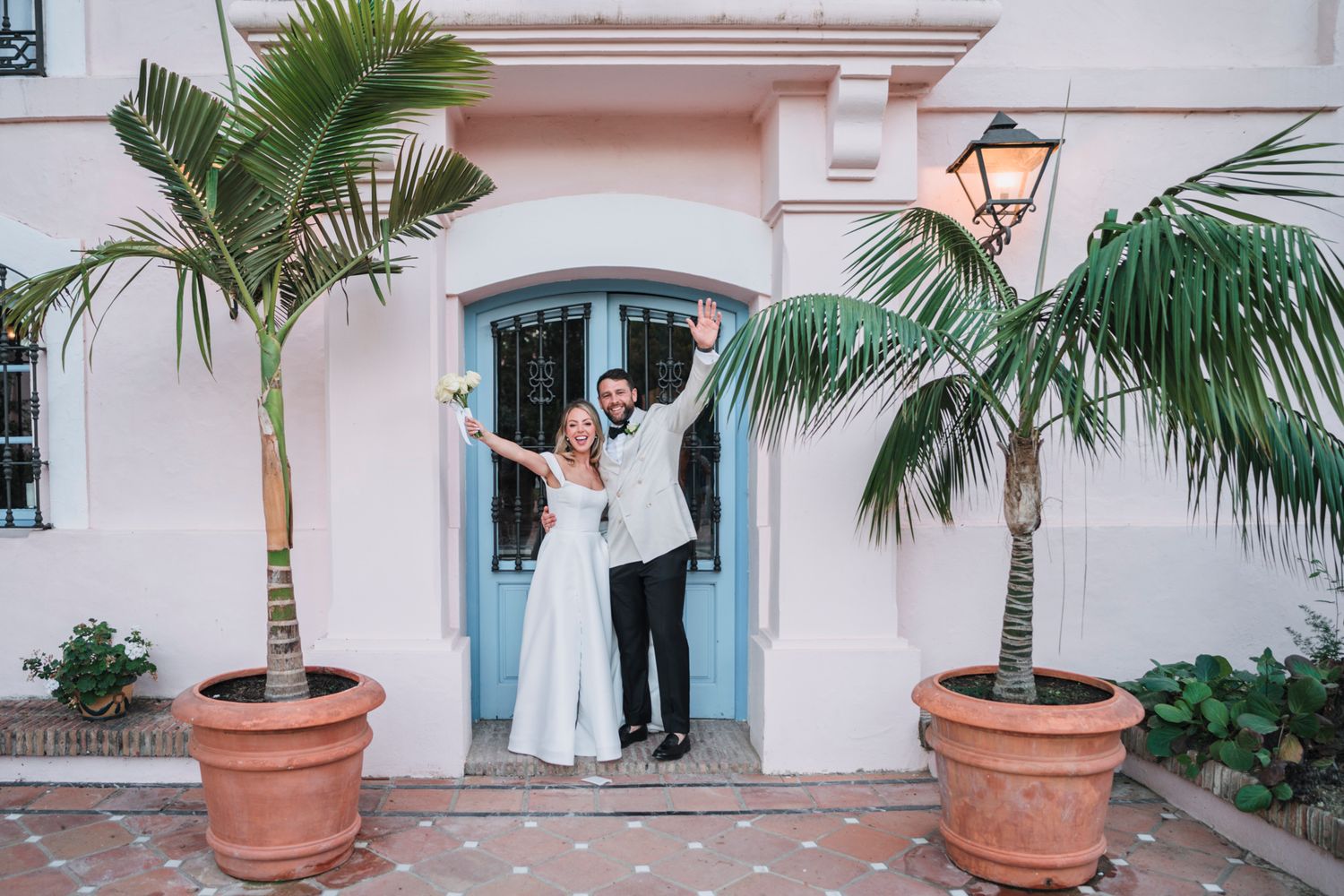 Two people stand between potted palm trees at the entrance of a pink stucco building with a blue door and vintage lamp.