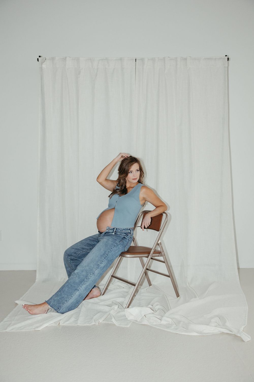 Person in denim overalls poses artistically with a chair against a white backdrop.