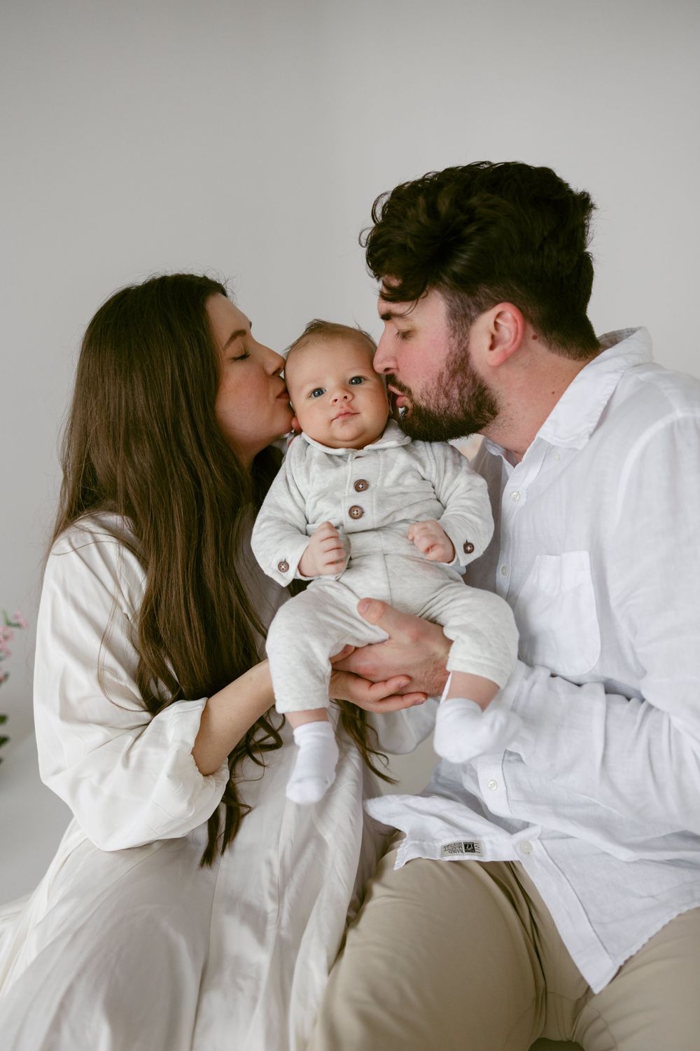 Family in white clothing sharing loving kisses with their baby in a minimalist setting.