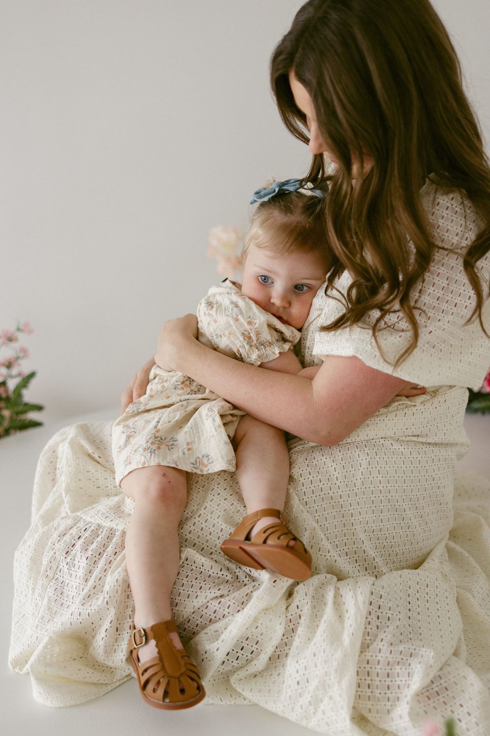 Mother in white lace dress holding toddler wearing matching outfit and brown sandals.