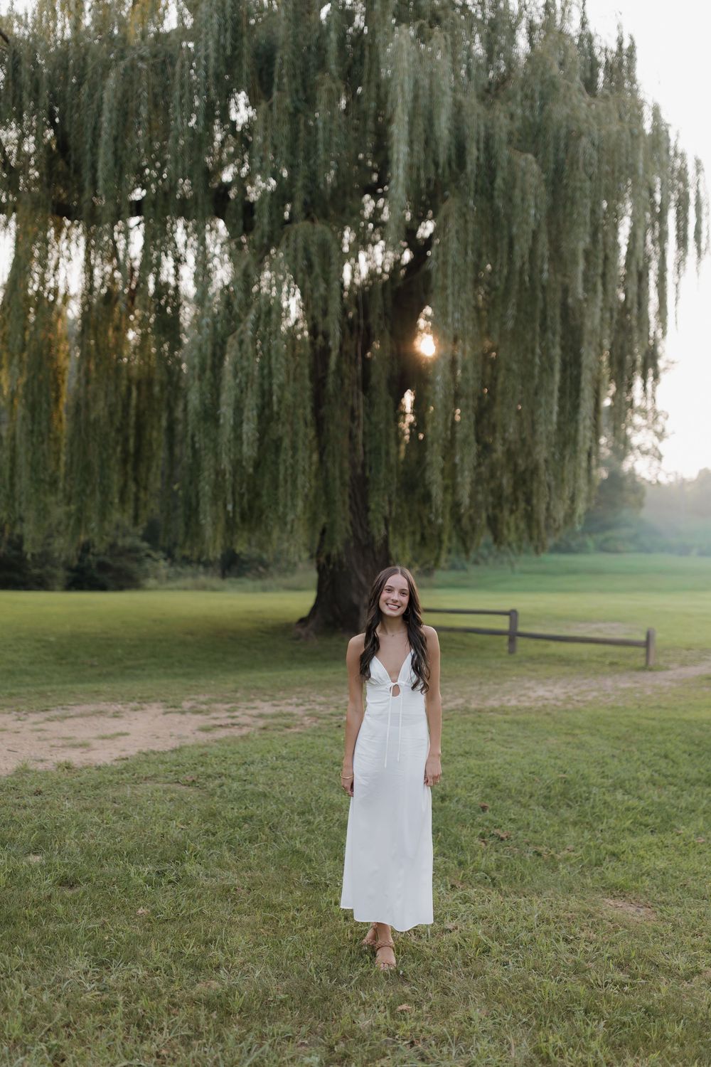 A figure in a white dress stands in a serene park setting with weeping willow trees at sunset.
