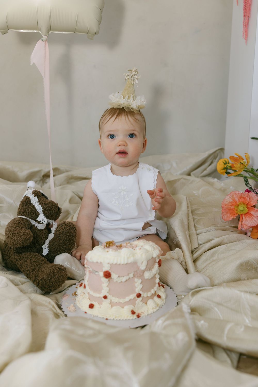Baby with birthday cake and teddy bear on cream bedding for first birthday celebration.