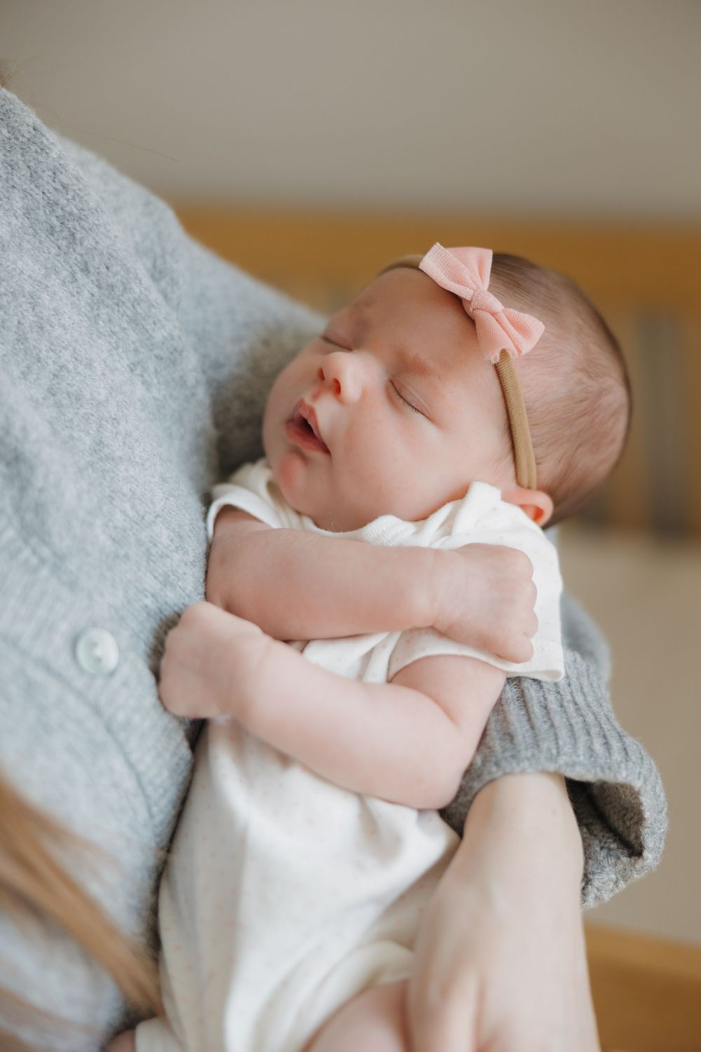 Sleeping newborn baby wearing pink bow headband and white onesie on gray blanket.