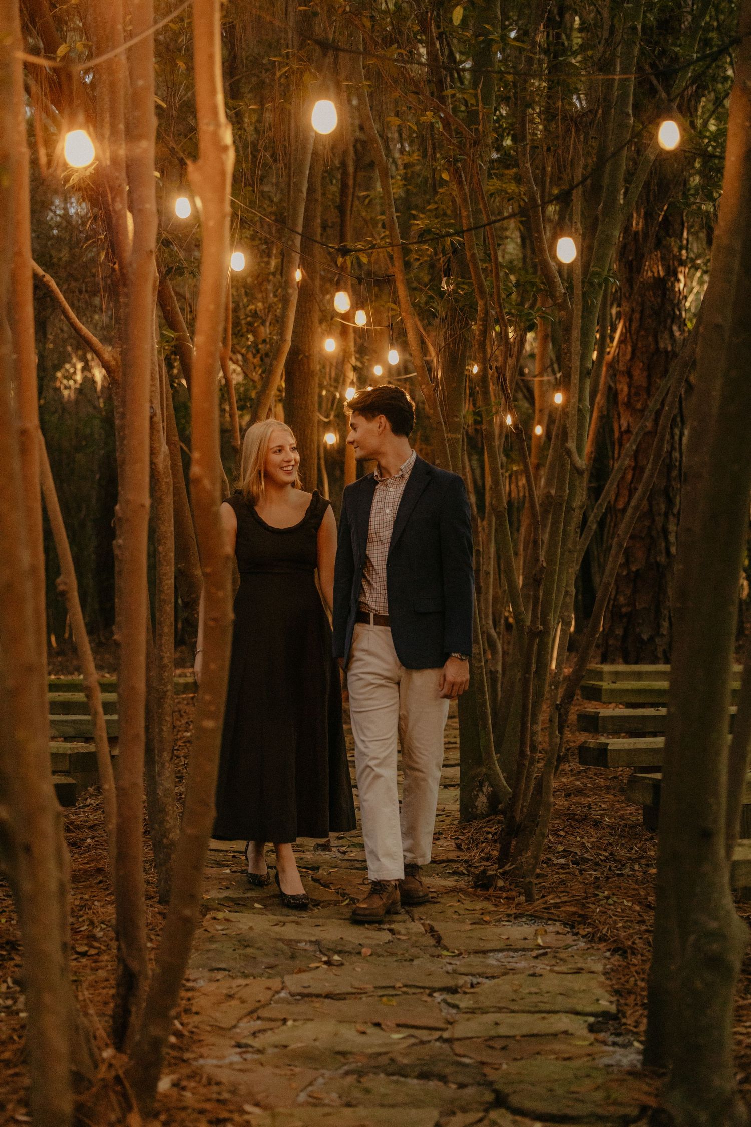 Couple walking hand in hand on a stone path surrounded by trees and warm string lights during golden hour session in Mobile, Alabama
