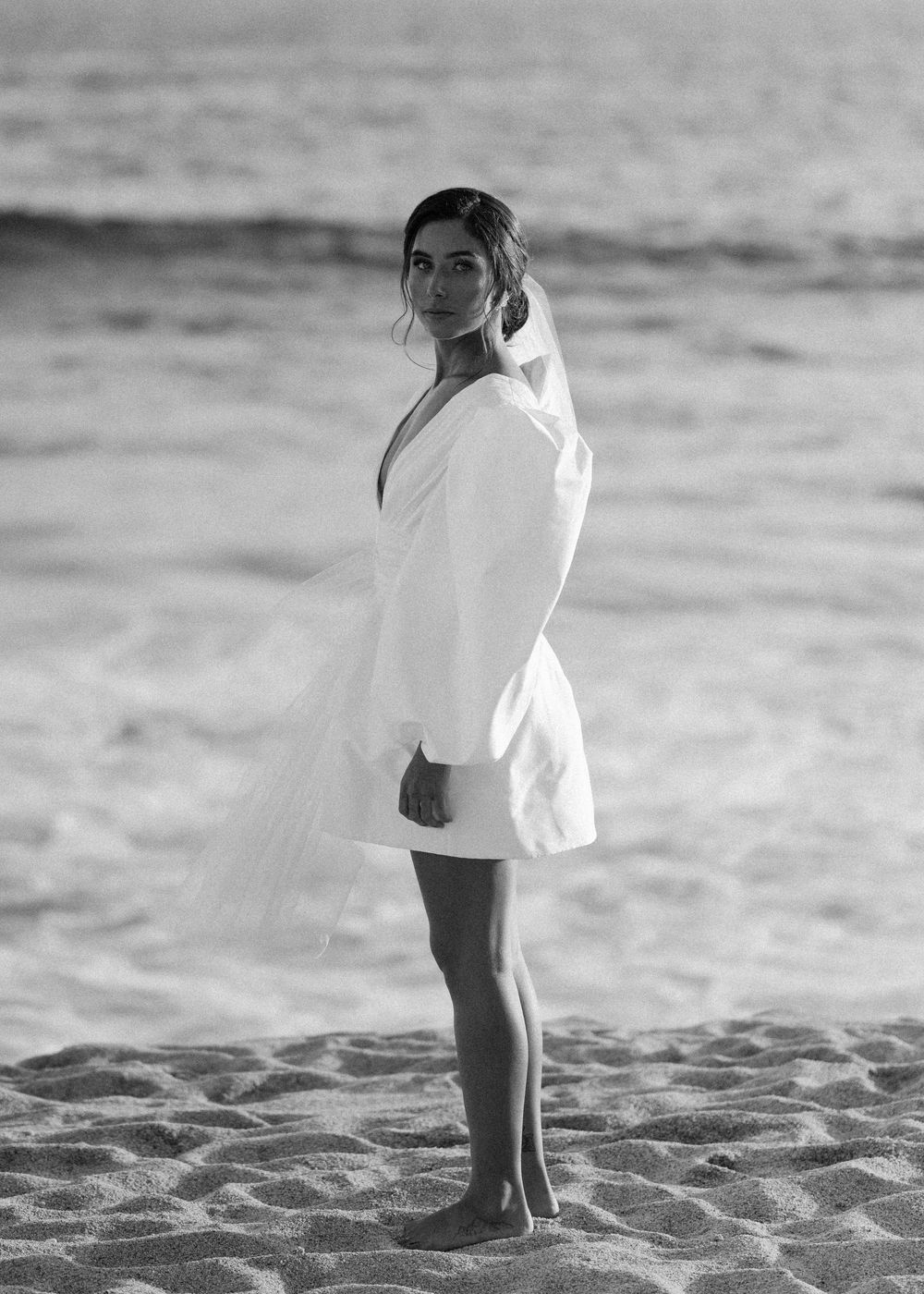 Woman in white dress poses on beach in artistic black and white portrait.