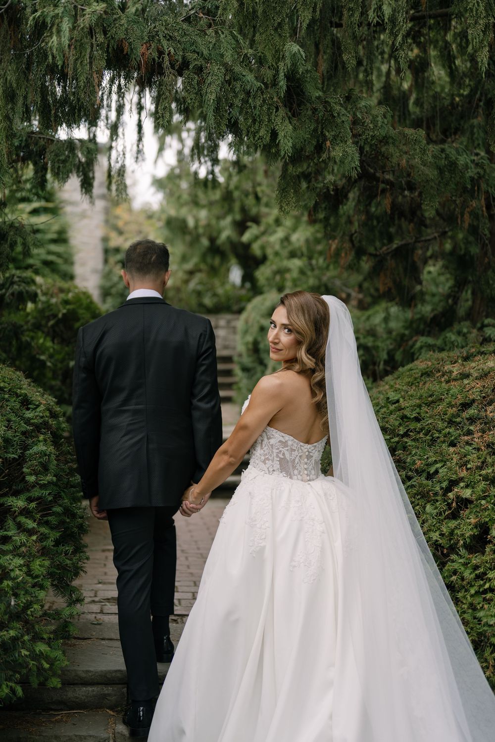 Bride and groom walk down garden path surrounded by lush greenery.