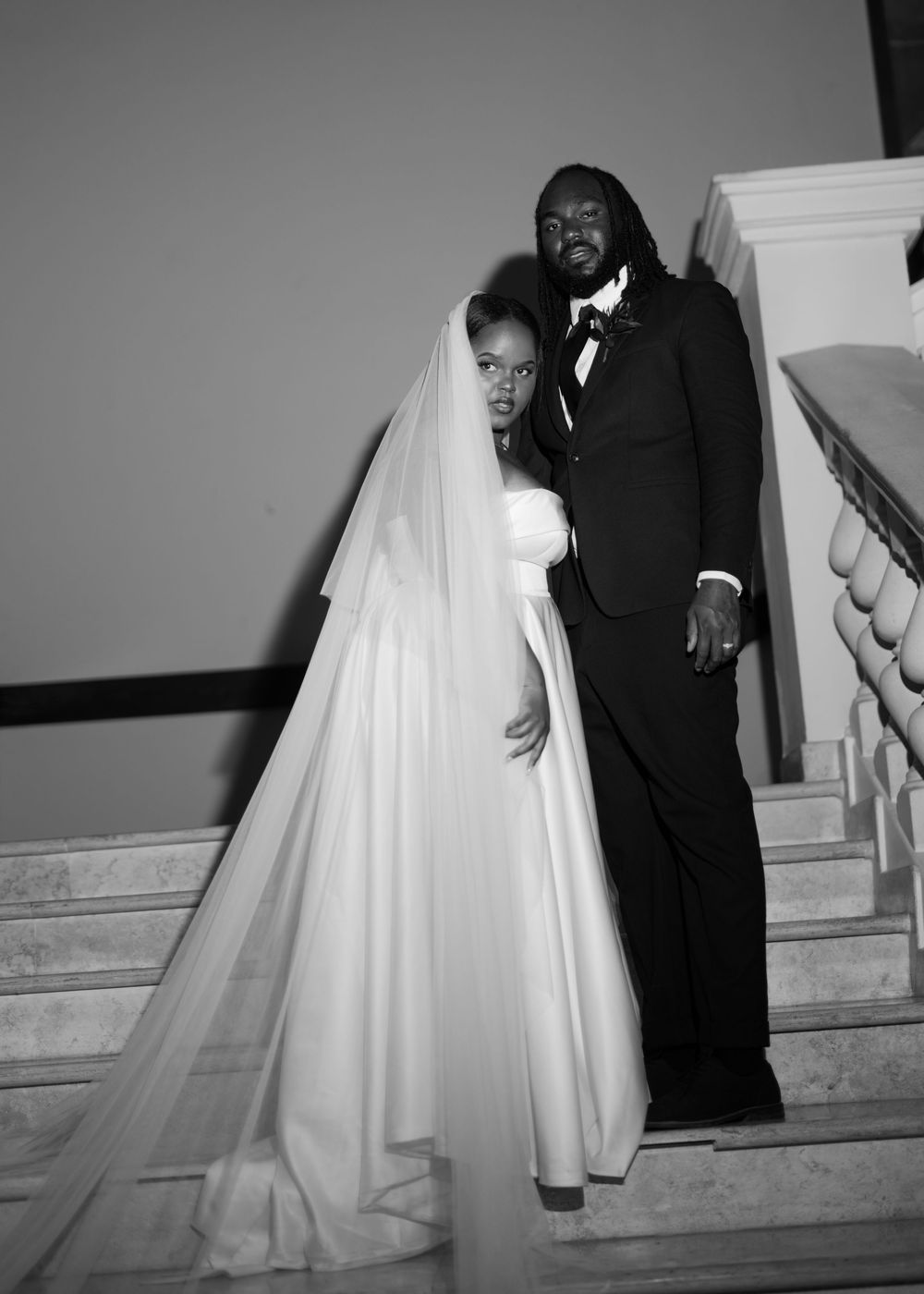 Wedding couple poses on grand staircase in dramatic black and white photography.