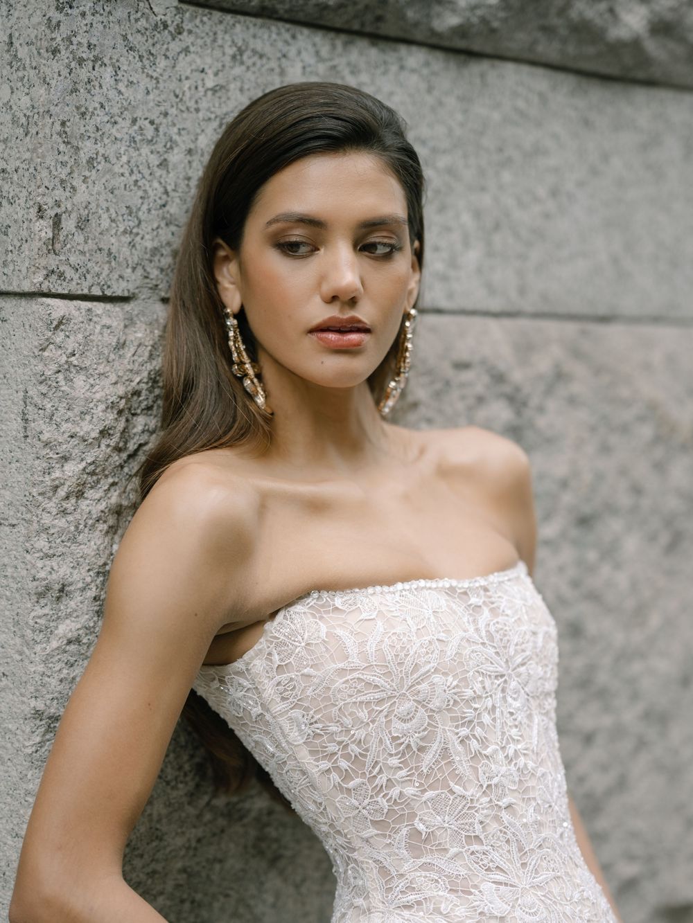 A close-up fashion photo of a strapless white lace wedding gown against a gray concrete wall background.