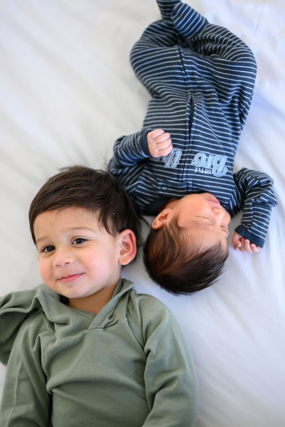 Two young children lying on white bedding, one wearing olive green and one in blue striped clothing.