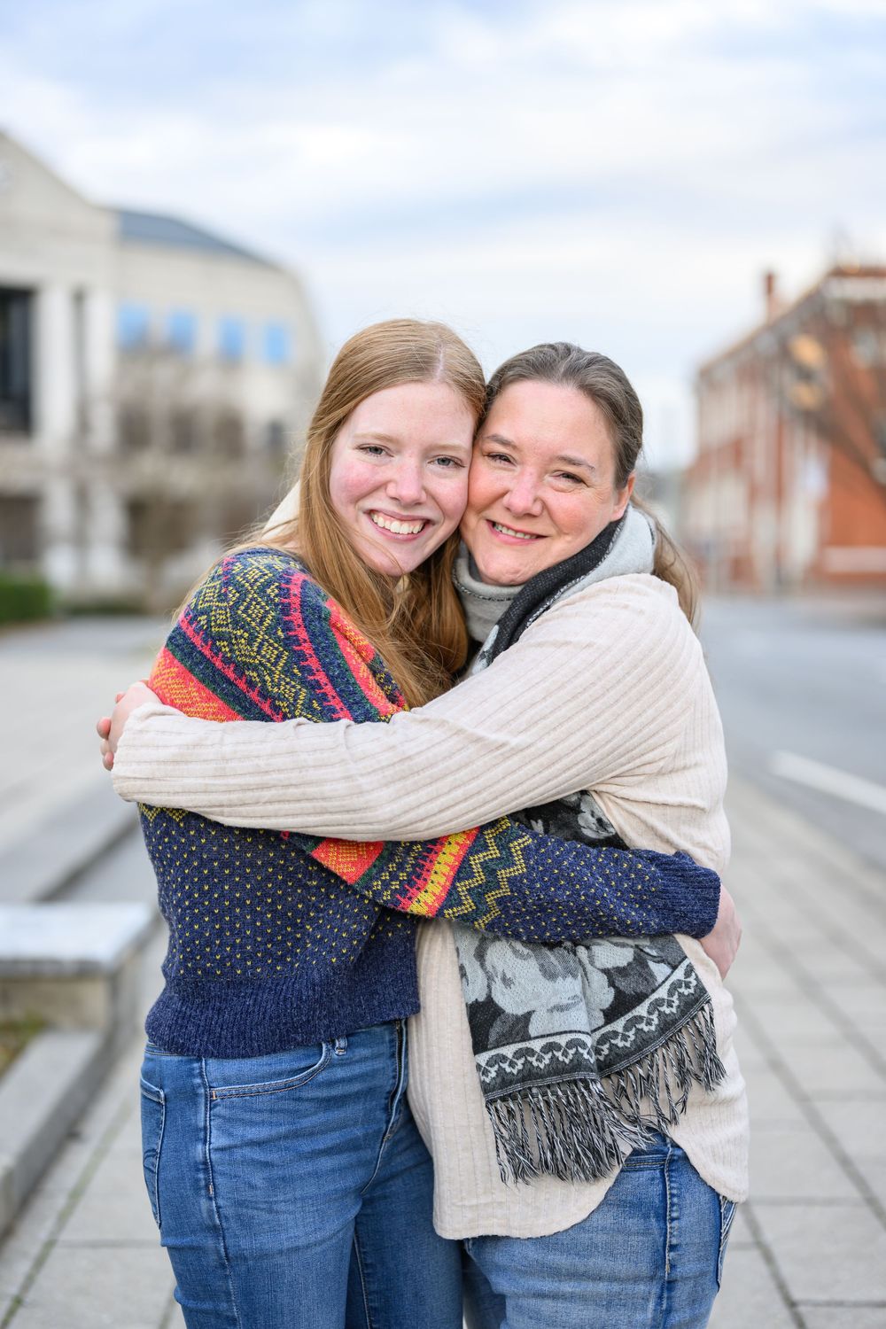 Two people in cozy sweaters embrace and smile on a city street during a chilly day.
