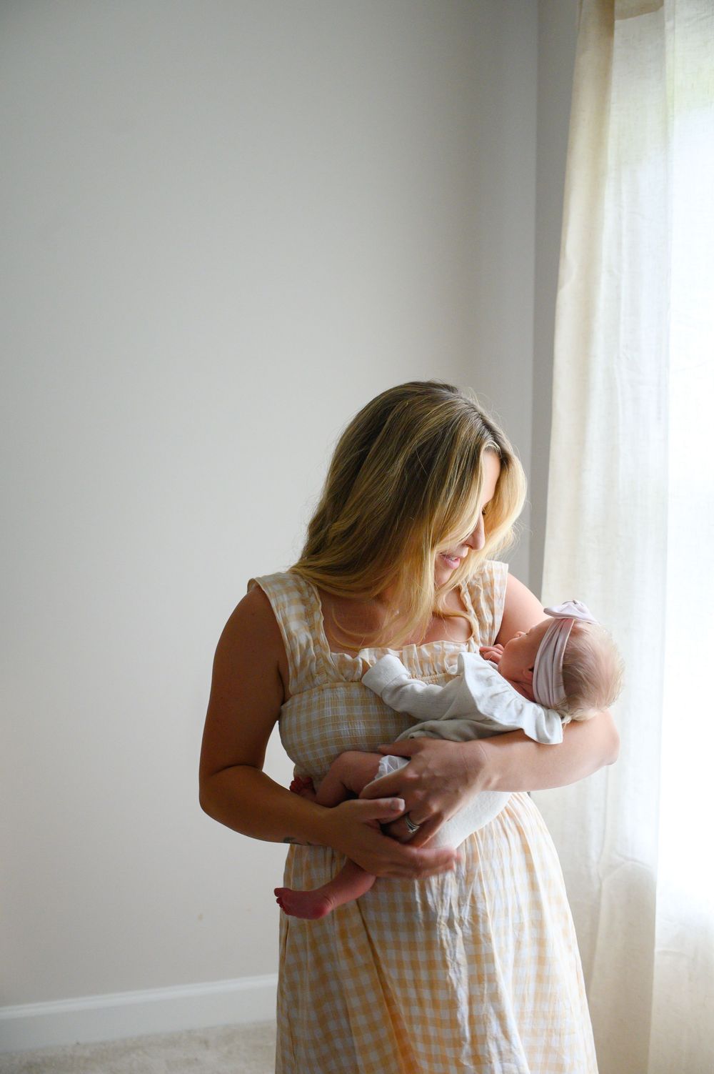 A tender moment by a bright window as someone in a checkered dress holds a swaddled newborn baby. family photos in canton, ga