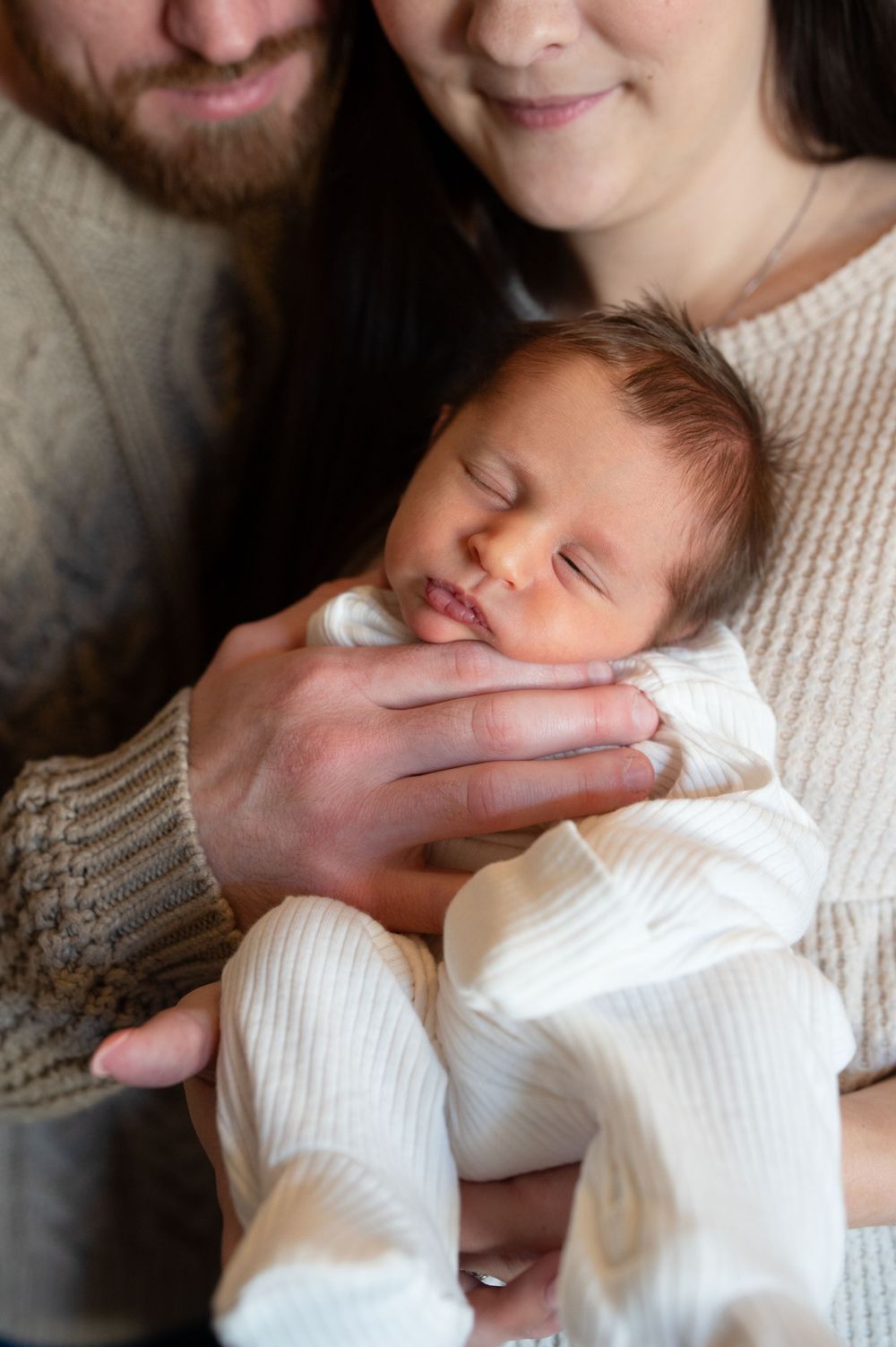 A sleeping newborn baby in white clothing is gently cradled by parents wearing cozy sweaters.