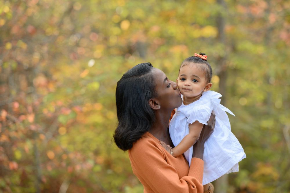 A loving adult wearing an orange sweater holds and kisses a small child in a white dress against autumn foliage.