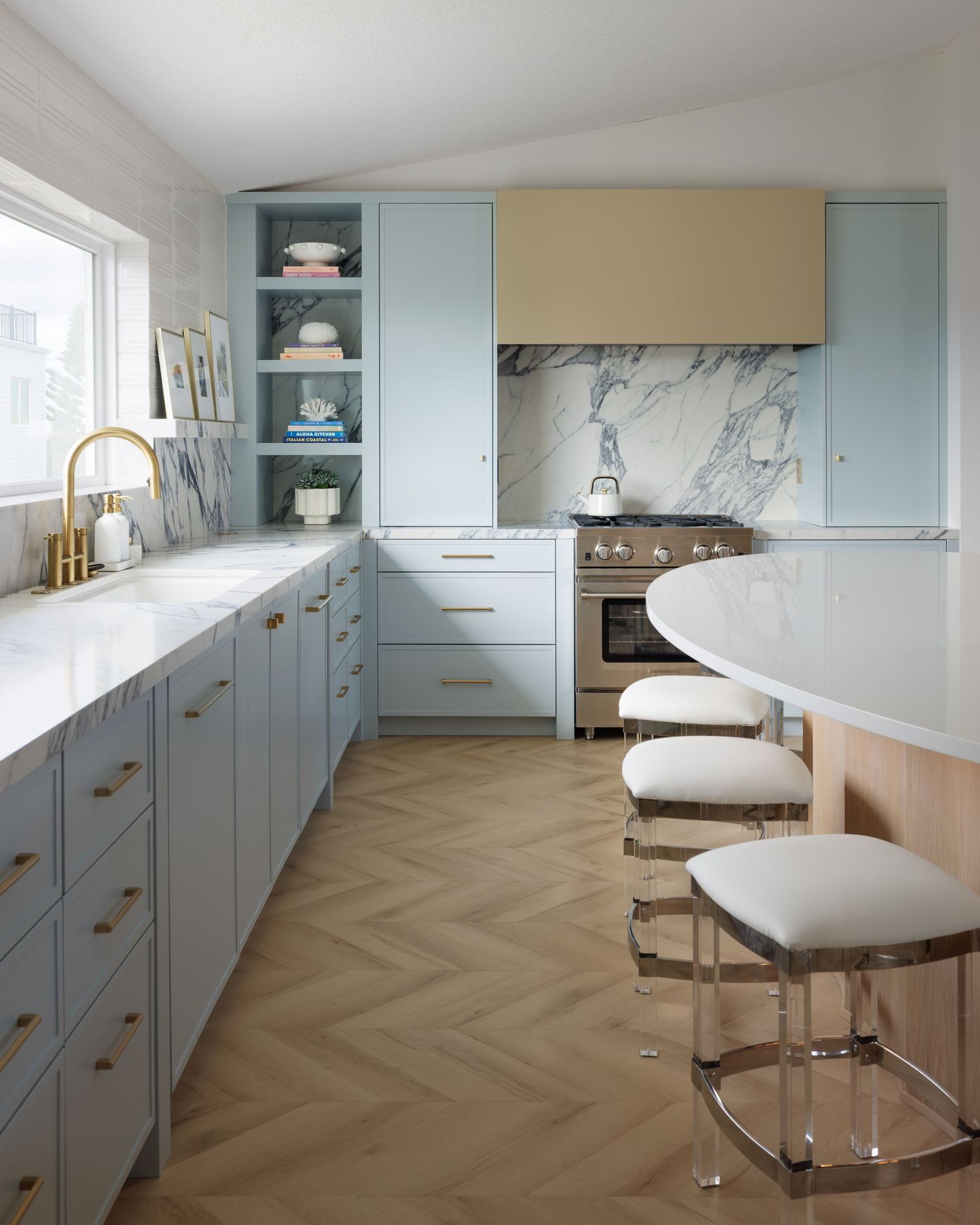 Light blue kitchen with brass hardware, marble backsplash, herringbone wood floors, and acrylic bar stools at island.