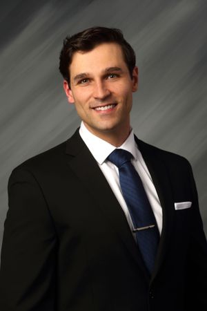 Professional headshot of someone in a dark suit with blue tie against a gray backdrop.