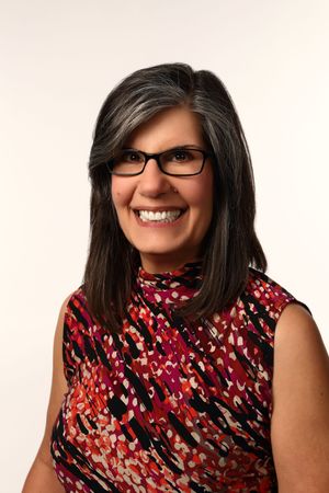 Smiling woman with dark hair and glasses wearing a colorful red patterned sleeveless top against white background.