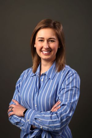Corporate portrait with crossed arms wearing blue striped shirt against dark backdrop.