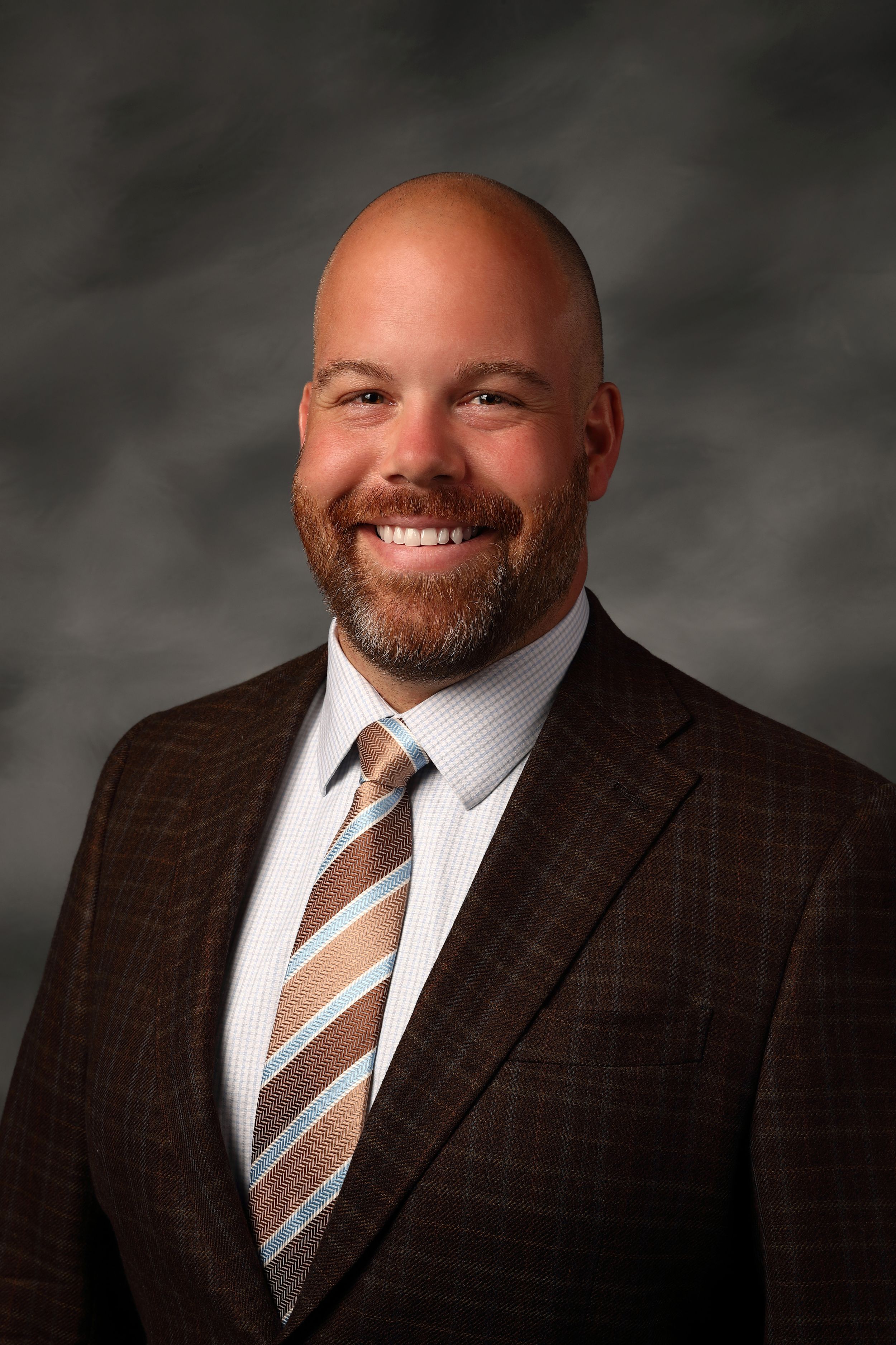 Professional portrait of person in brown plaid suit jacket and striped tie against dark background styled studio lighting.