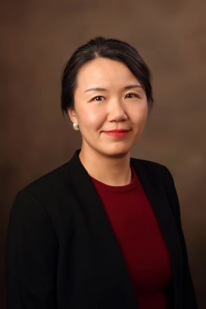 Professional headshot of person in black blazer and red top against brown background.