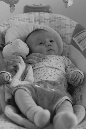 Black and white photo of a newborn baby lying in a bouncer seat holding a small stuffed animal toy.