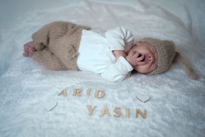 Newborn baby named Ario Yasin lying on white fur blanket wearing beige knit outfit with wooden letter name display.
