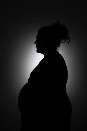 Silhouette of a pregnant woman in profile against a dark background with soft backlight highlighting her figure.