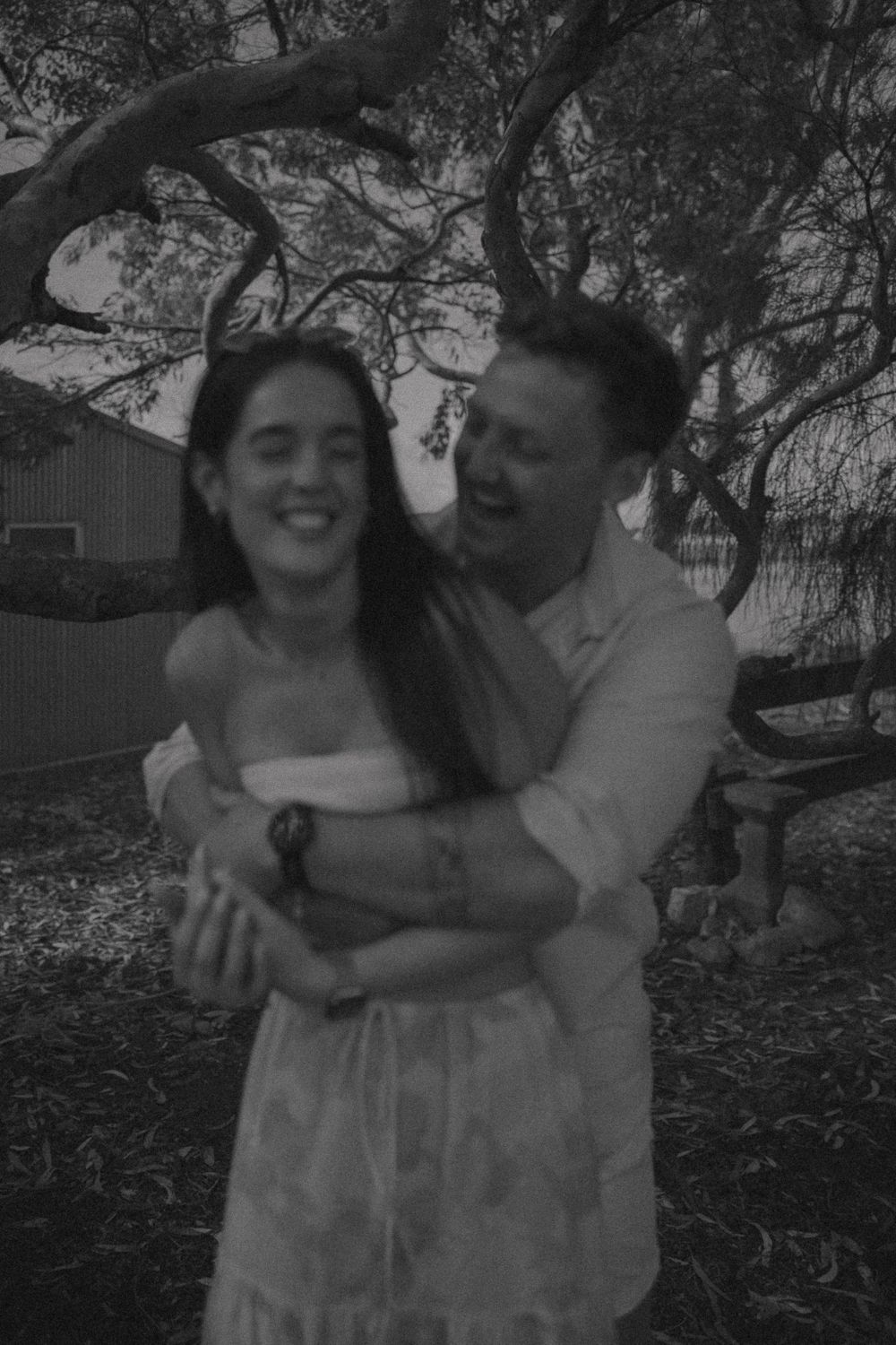 A joyful black and white photo captures a playful moment between two people in a wooded setting.