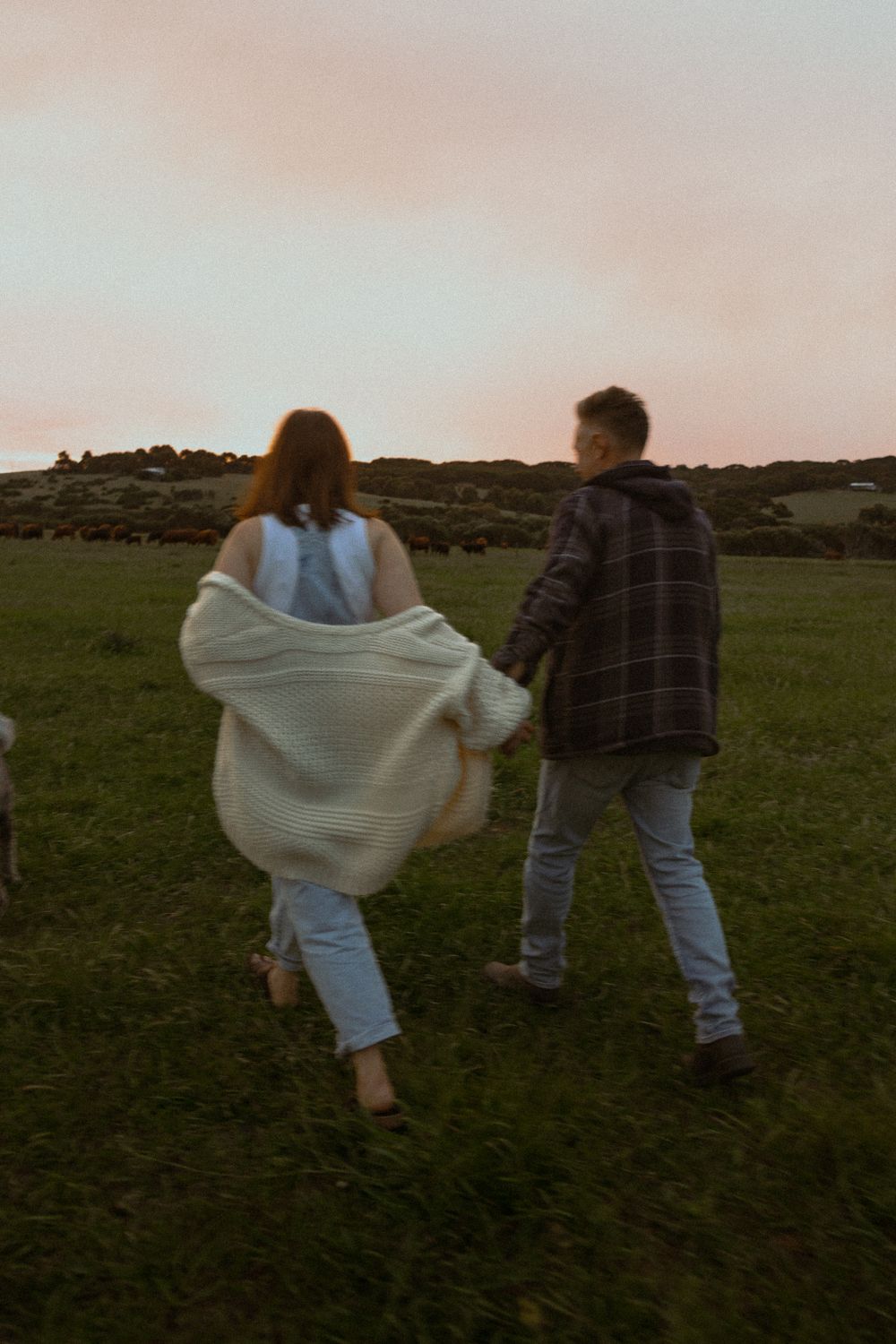 A couple walks through a grassy field at dusk with flowing fabric catching the wind.