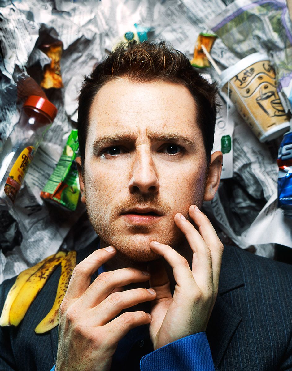 A young red-haired man in a suit poses thoughtfully surrounded by crumpled trash and garbage.