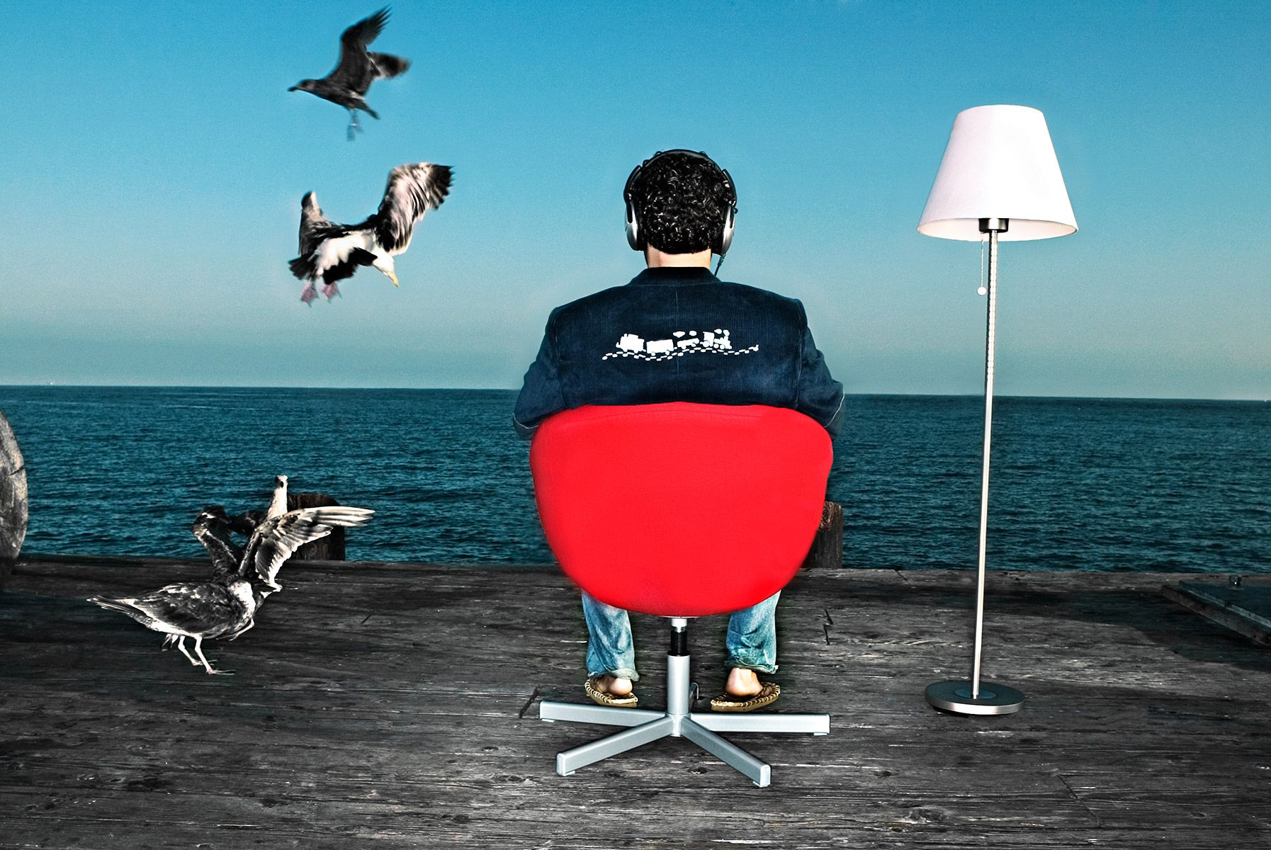 A person sits in a red chair facing the ocean while birds and a dog fly around them.
