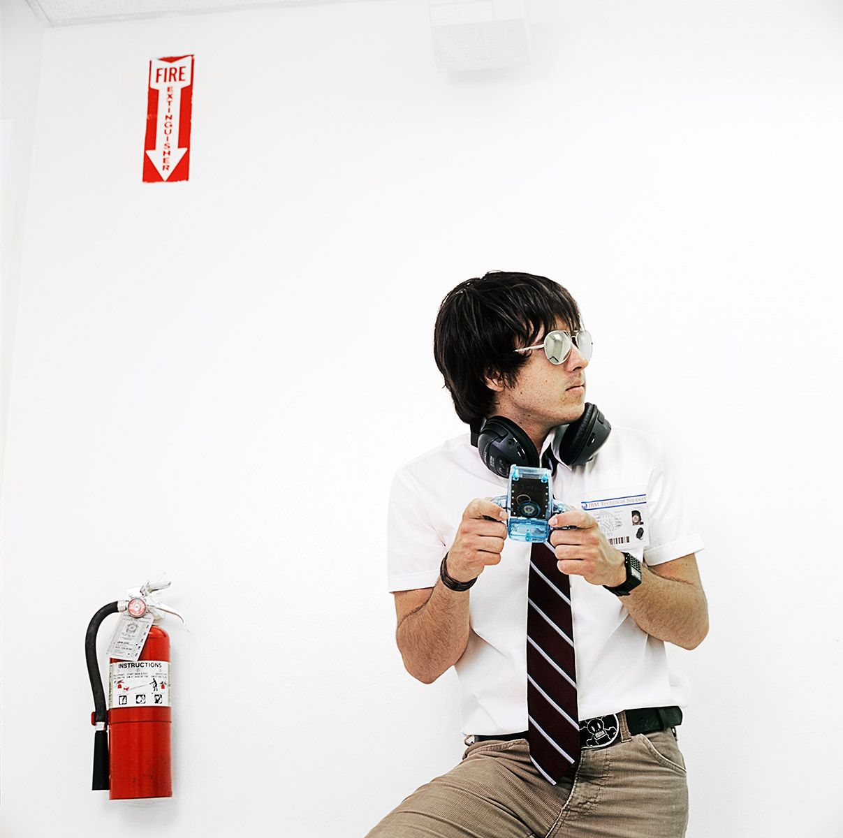 A young Asian man in a white shirt and tie holds an ID badge near a fire extinguisher.