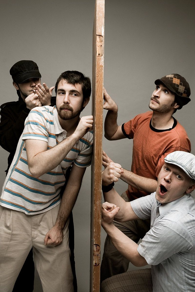 Four young men in casual clothes dramatically pose around a wooden pole against a gray background.