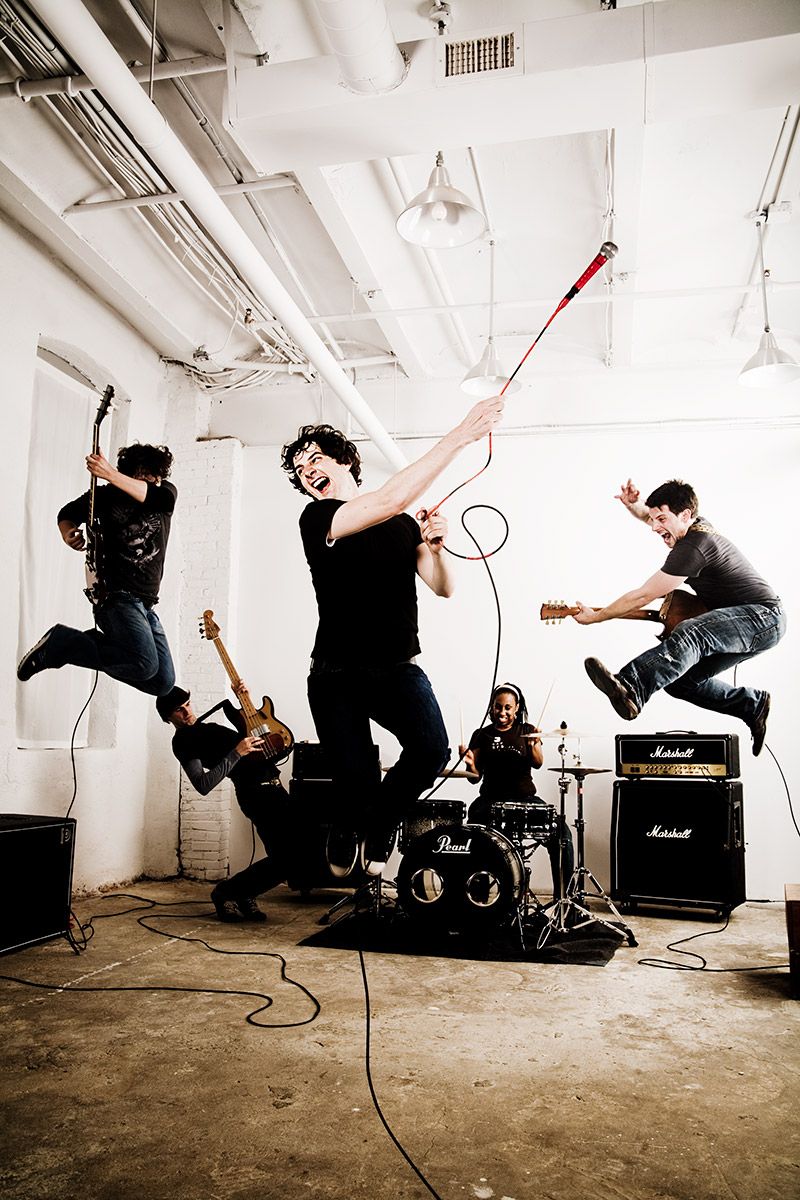 An energetic rock band jumps in the air while performing in a white industrial rehearsal space.