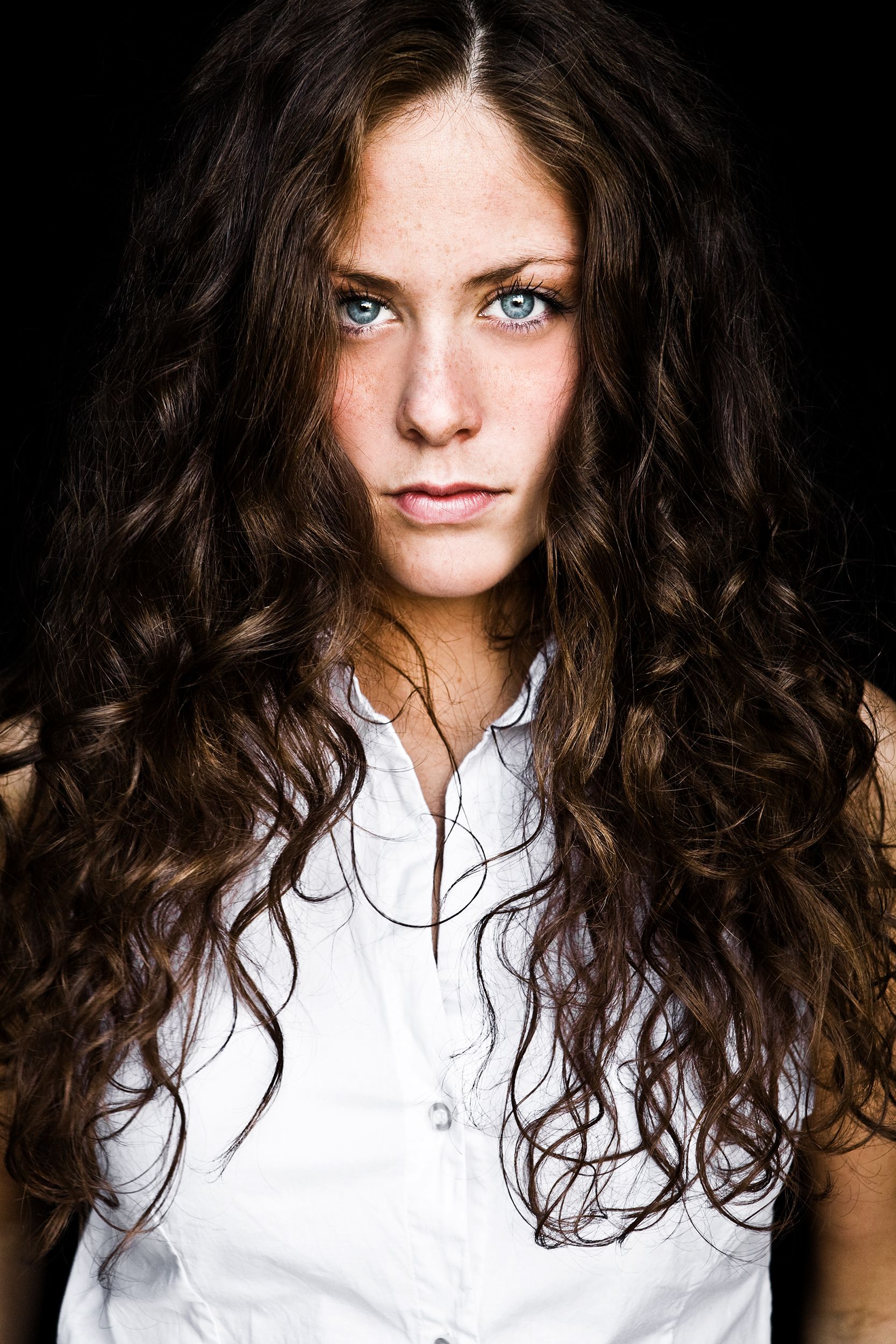 Young woman with long curly brown hair and blue eyes in white shirt on dark background.
