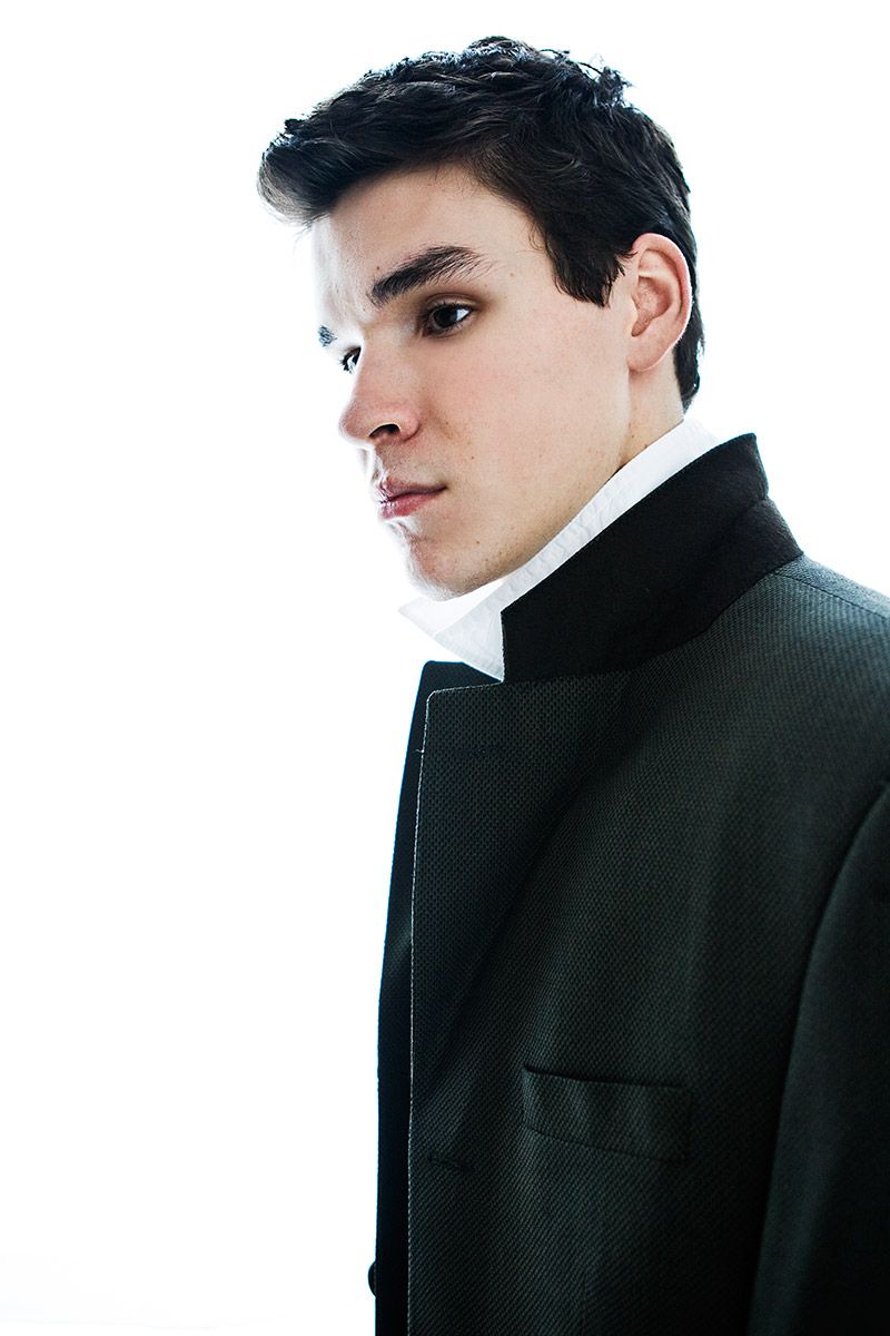 Young man in black tuxedo jacket and white dress shirt posing against white background.